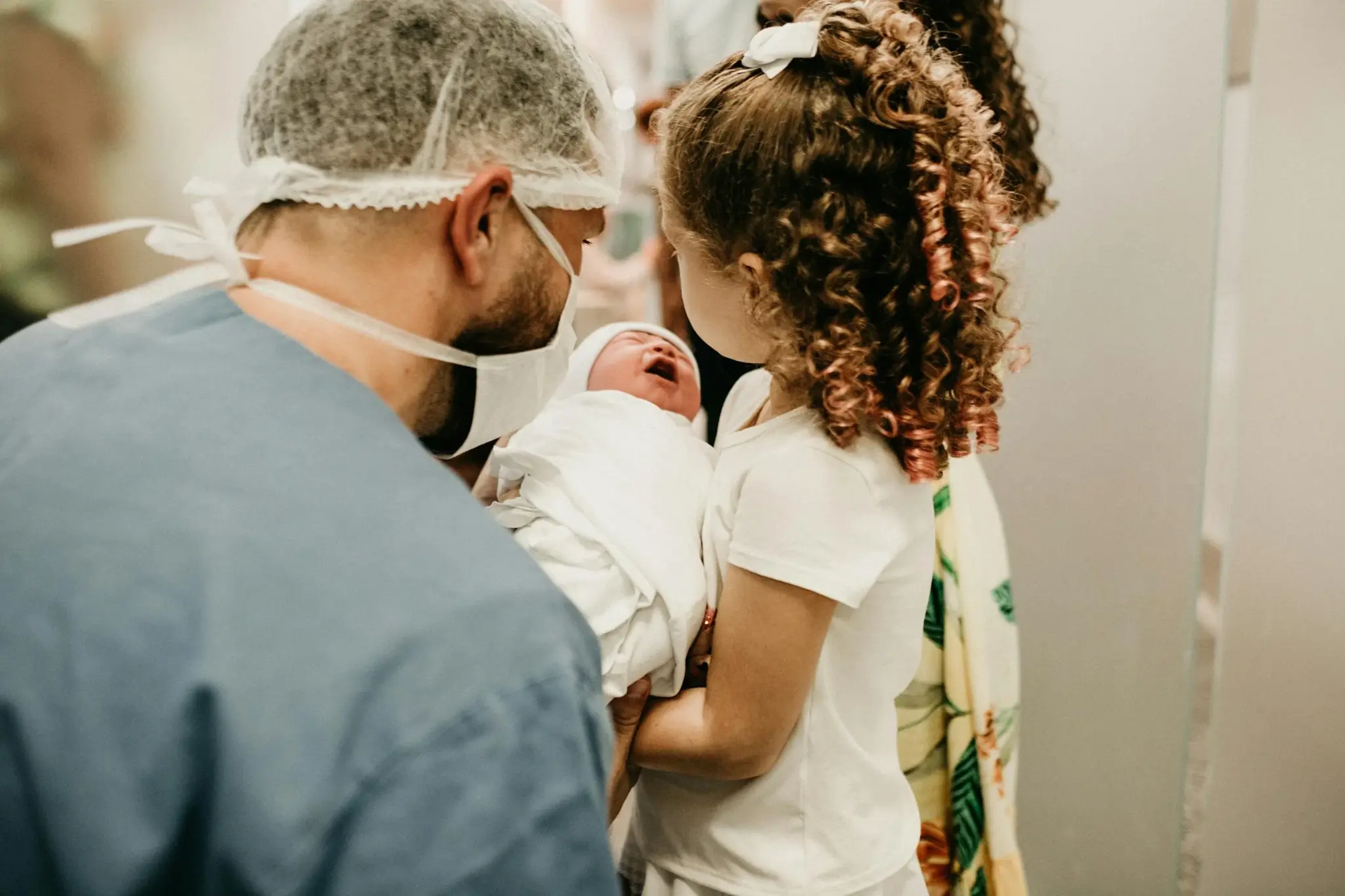 Pai com máscara cirúrgica segurando recém-nascido enquanto irmã mais velha observa o bebê em ambiente hospitalar
