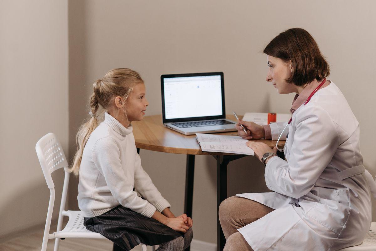 Médica conversando e avaliando uma menina sentada à mesa durante consulta pediátrica