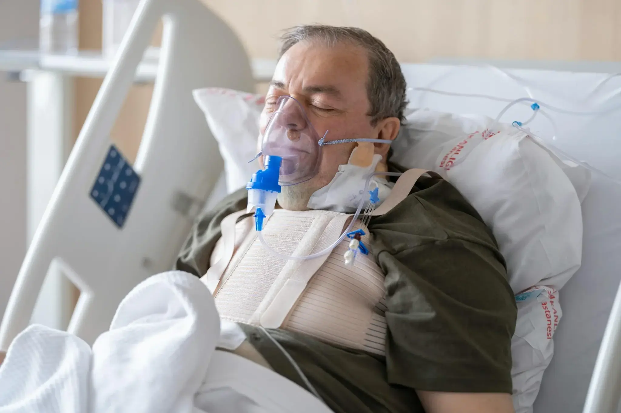 Paciente em leito hospitalar utilizando máscara de oxigênio, com colar cervical e cateteres visíveis