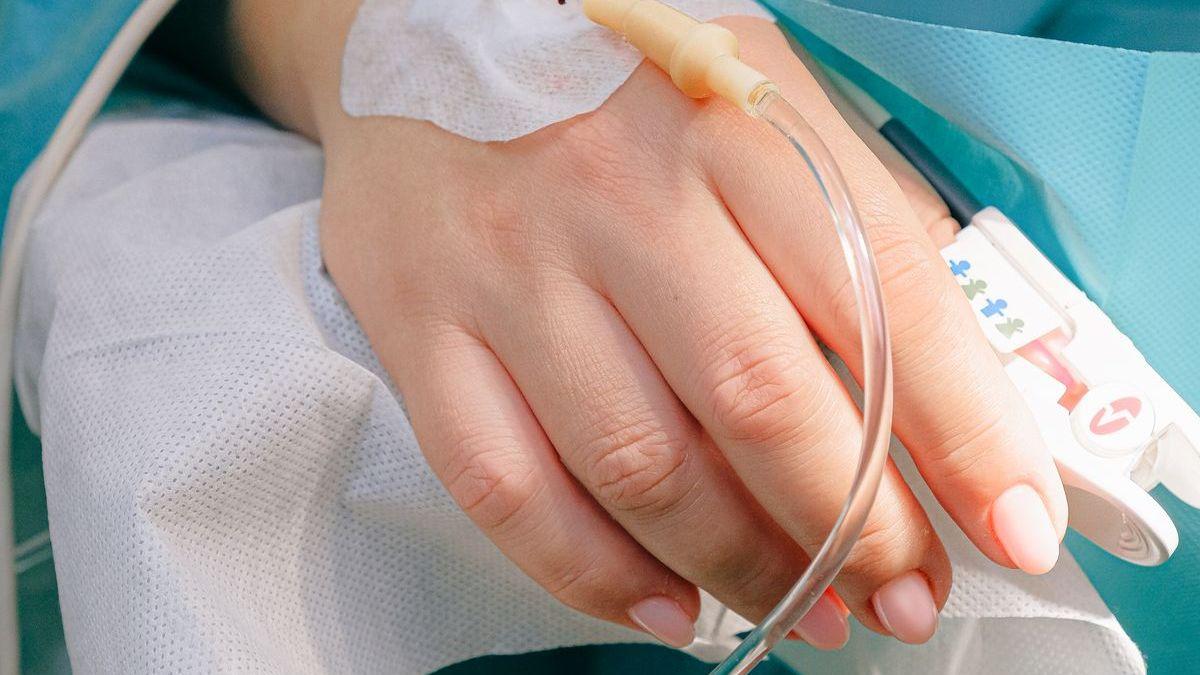 Mãos com luvas cirúrgicas segurando cateter de acesso vascular fixado no dorso da mão de um paciente