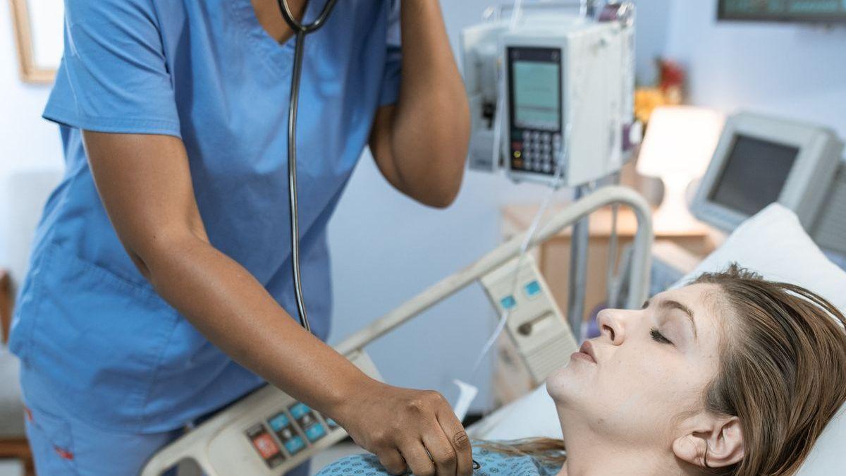 Enfermeira com máscara auscultando o tórax de paciente internado, cercada de bombas de infusão e monitor multiparâmetro