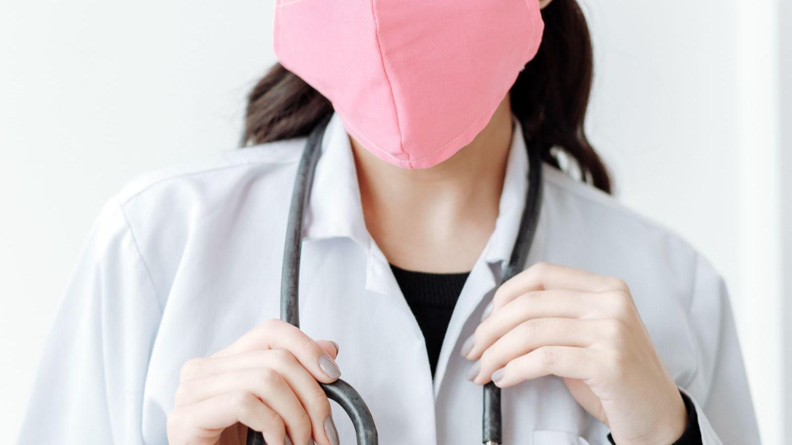 Médica com máscara e jaleco branco segurando o estetoscópio junto ao pescoço, pronta para atendimento de emergência