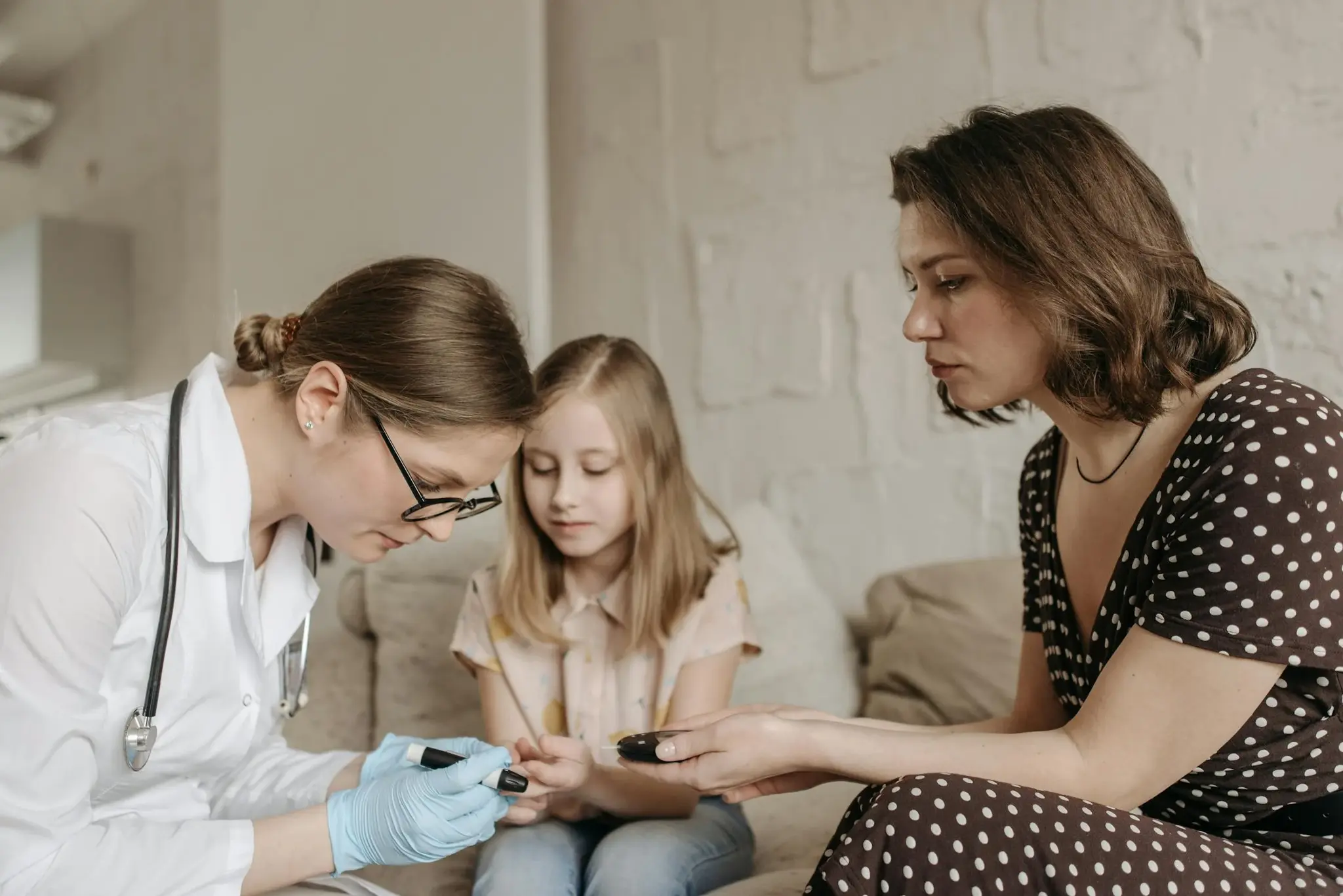 Médica realizando teste de glicemia capilar no dedo de uma menina, com a mãe observando ao lado