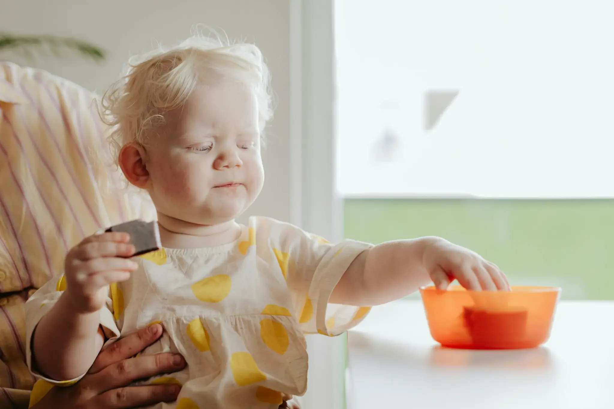 Bebê loiro sentado à mesa alcançando uma tigela laranja com pedaços de comida